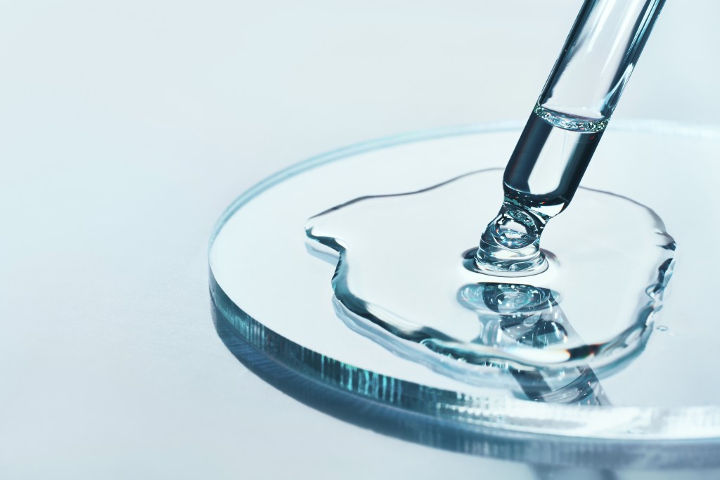 Pipette with sample of cosmetic product in petri dish on blue background, selective focus.