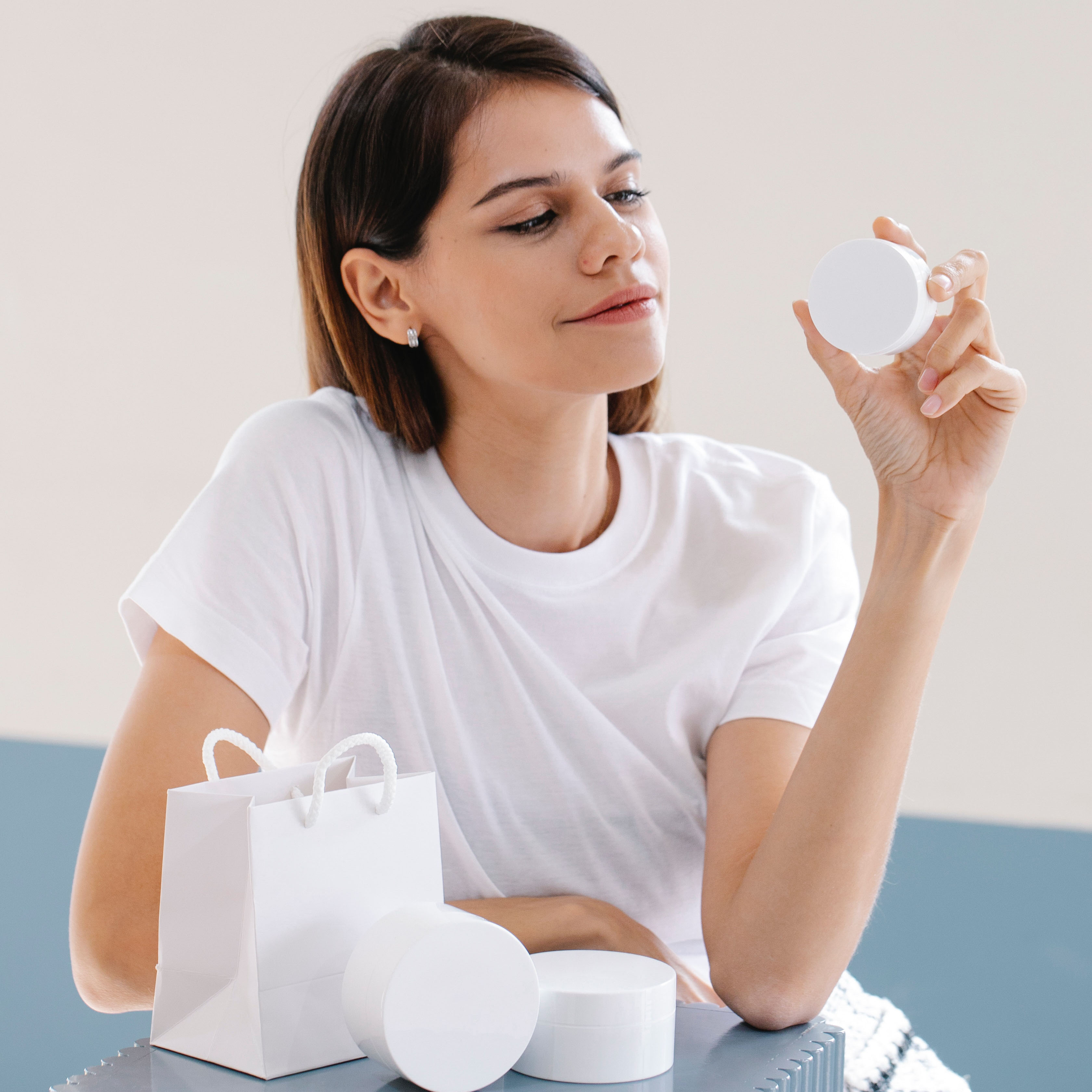 Woman sitting and holding jar of unlabeled skincare product, admiring it, while other jars are right next to her on a table